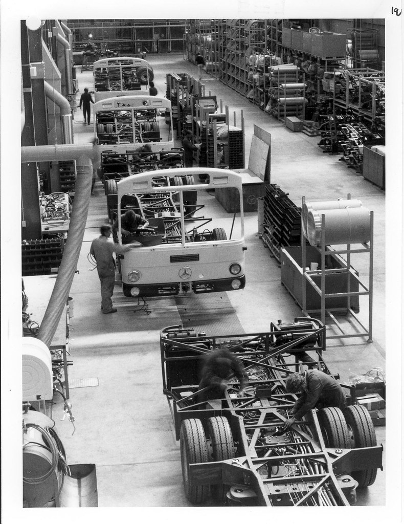 Workers assembling Mercedes-Benz buses on a factory floor, with exposed chassis and partly completed bus bodies.