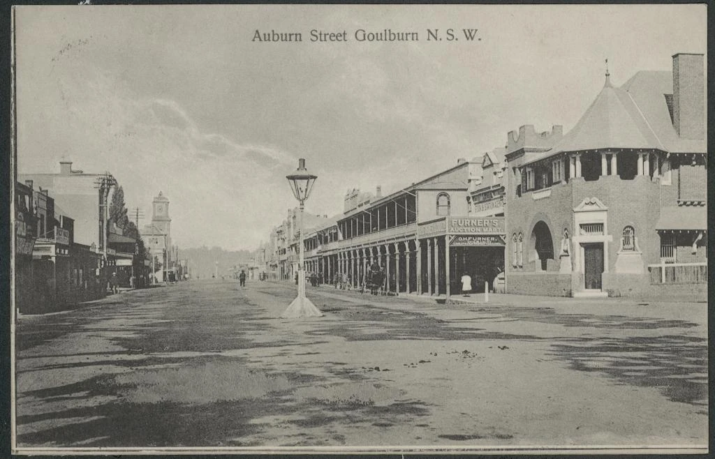 Historical postcard showing Auburn Street in Goulburn, with a central street lamp, shop verandas and a distant clock tower.