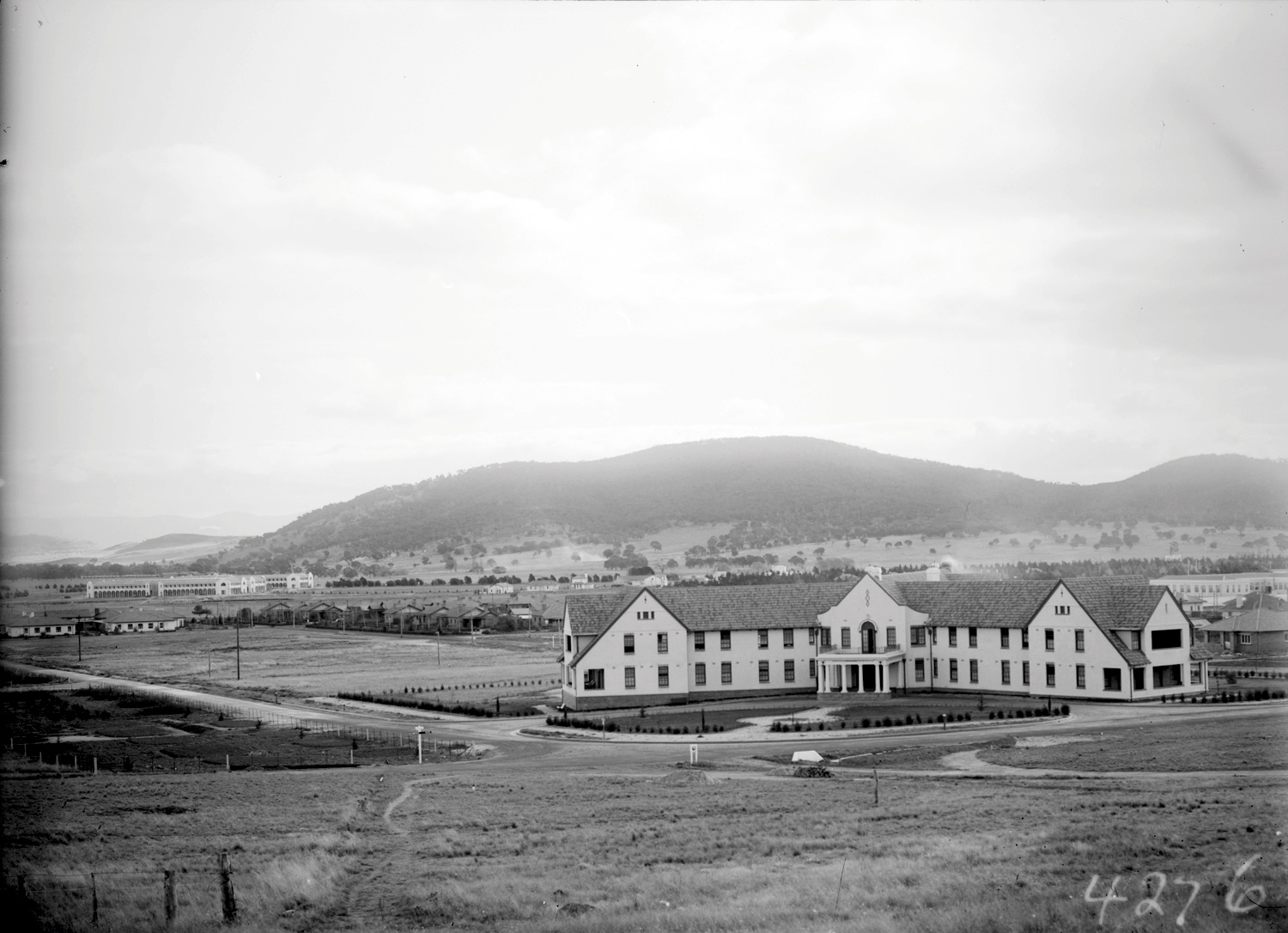 Hotel Ainslie’s pale walls and tiled roofs beyond open ground, with scattered buildings and hills in the distance.