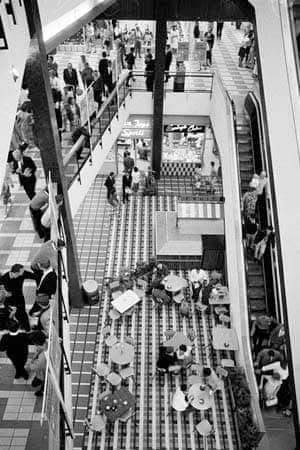 Monaro Mall seen from an upper level, with escalators, shoppers and tables on the tiled floor below.